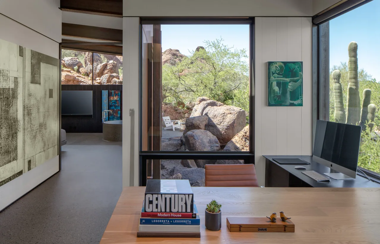 Home office with panoramic desert views including rocks and tall cacti seen through large black-framed windows.