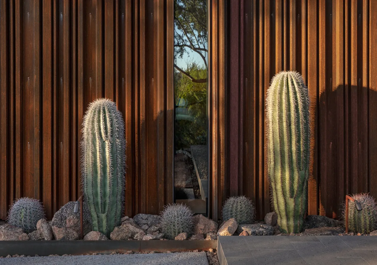 Rust-colored metal cladding with integrated desert rock landscaping and tall cacti in front of the exterior wall.
