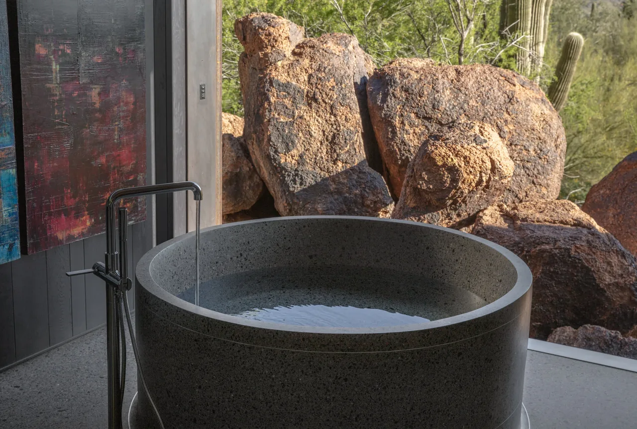 Round stone soaking tub with a black floor-mounted faucet next to a large window showing desert boulders.