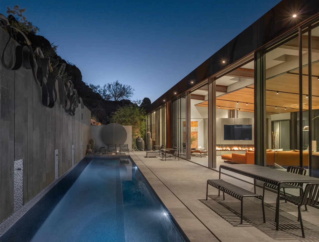 This modern home features floor-to-ceiling glass walls adjacent to a long pool and an outdoor dining area.