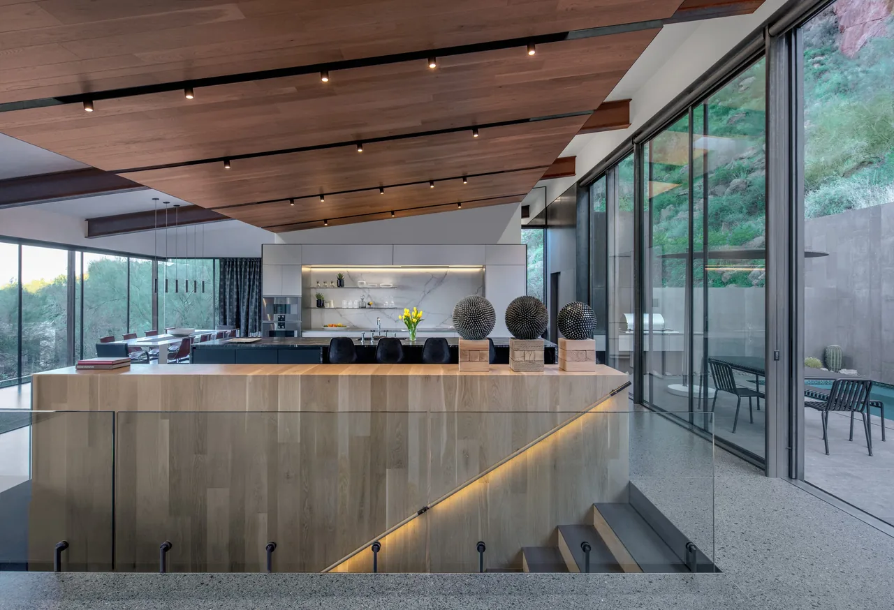 Open kitchen and dining space with wood ceiling, floor-to-ceiling windows overlooking desert rock and outdoor patio pool area.