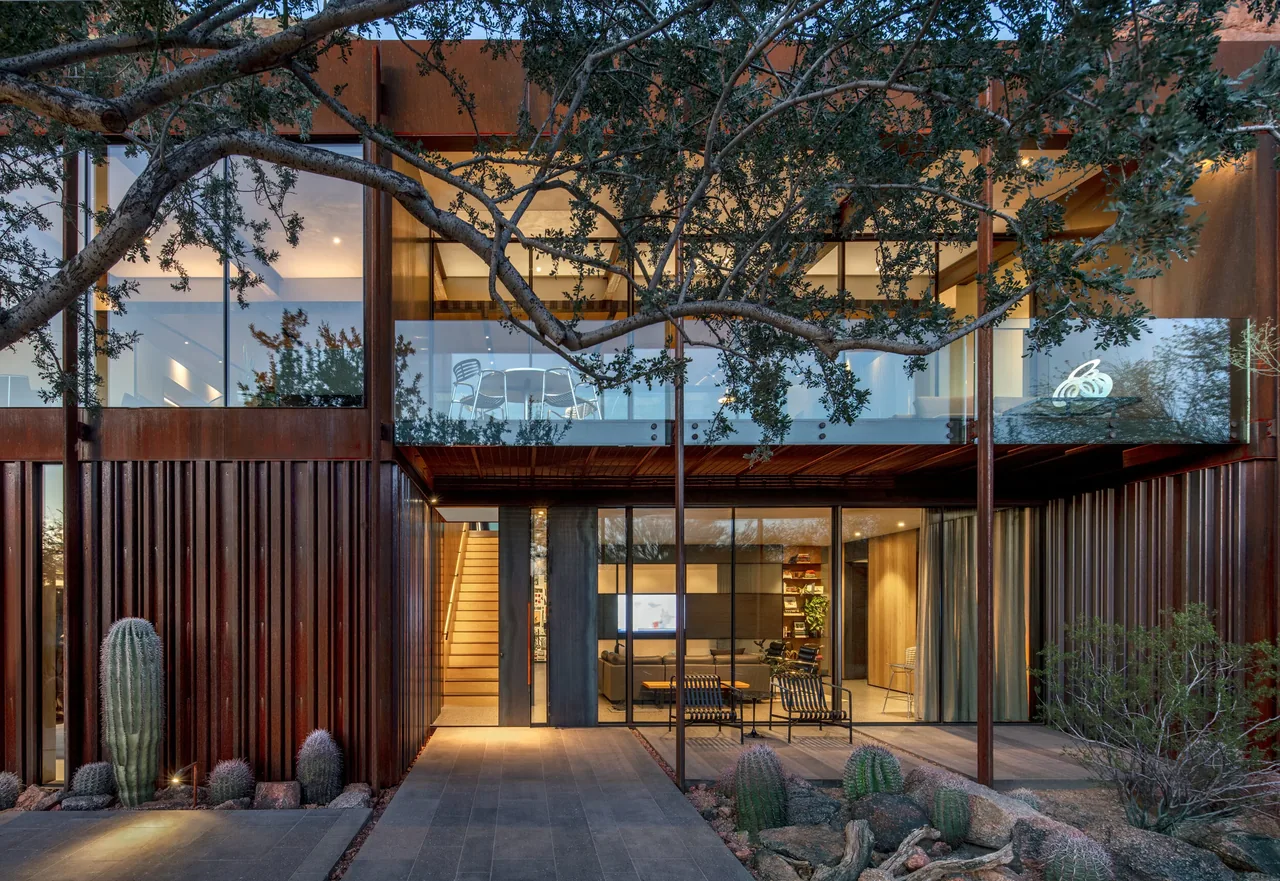Modern two-story home with weathered steel cladding and desert rock landscaping including tall cacti and outdoor seating on the patio.
