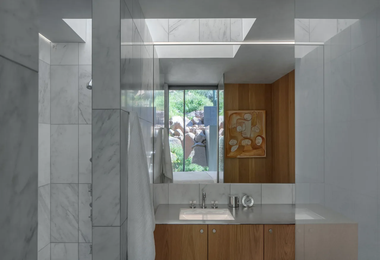 Interior view of a bathroom featuring marble wall tiles, a wood vanity, and a window showing desert rock landscape.