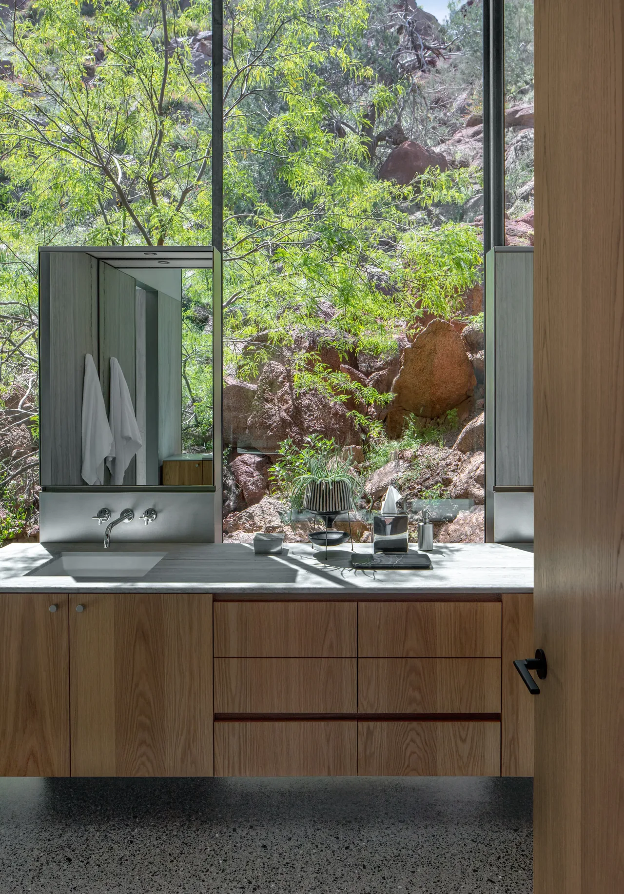 Bathroom vanity in wood with a large window framing desert rock and bright green foliage outside.