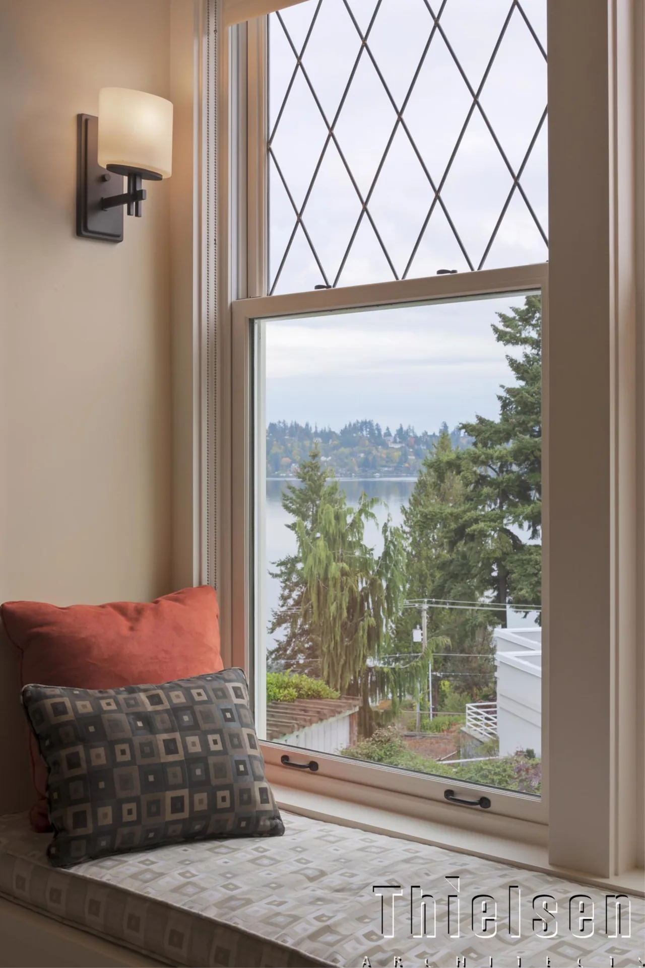 Window seat with patterned cushions beside a diamond-pane window overlooking a water and tree landscape.