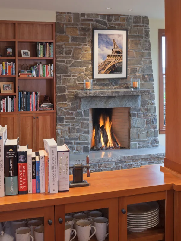 Interior view of a living room with a stone fireplace flanked by wood shelves and a wood cabinet displaying books and ceramics.