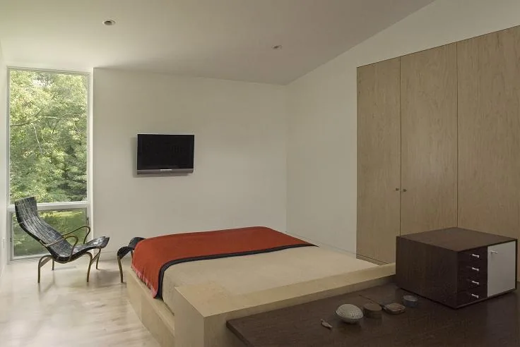 Ceruti House bedroom features minimalistic design with natural wood elements and large window overlooking green foliage.