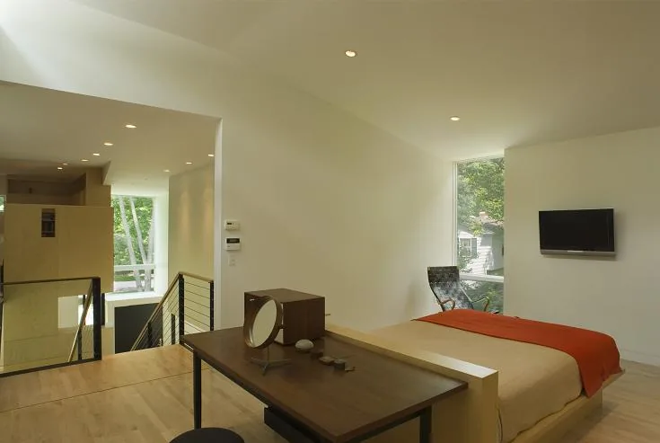 The bedroom features a minimalist design with a wooden desk, beige bedding, an orange throw, and a large narrow window.