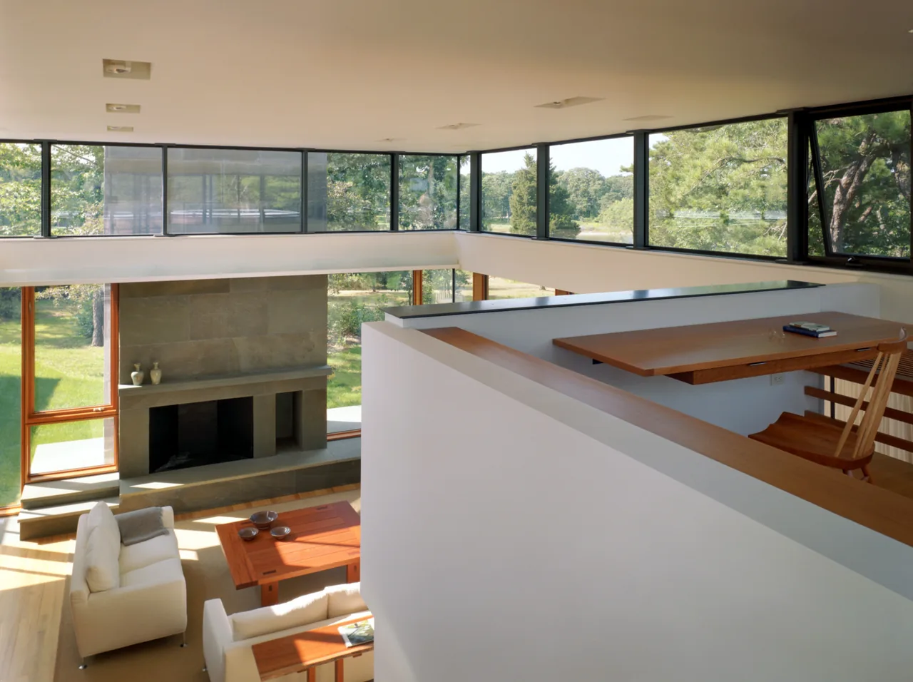 Two-story living room with stone fireplace and upper loft workspace surrounded by large windows.