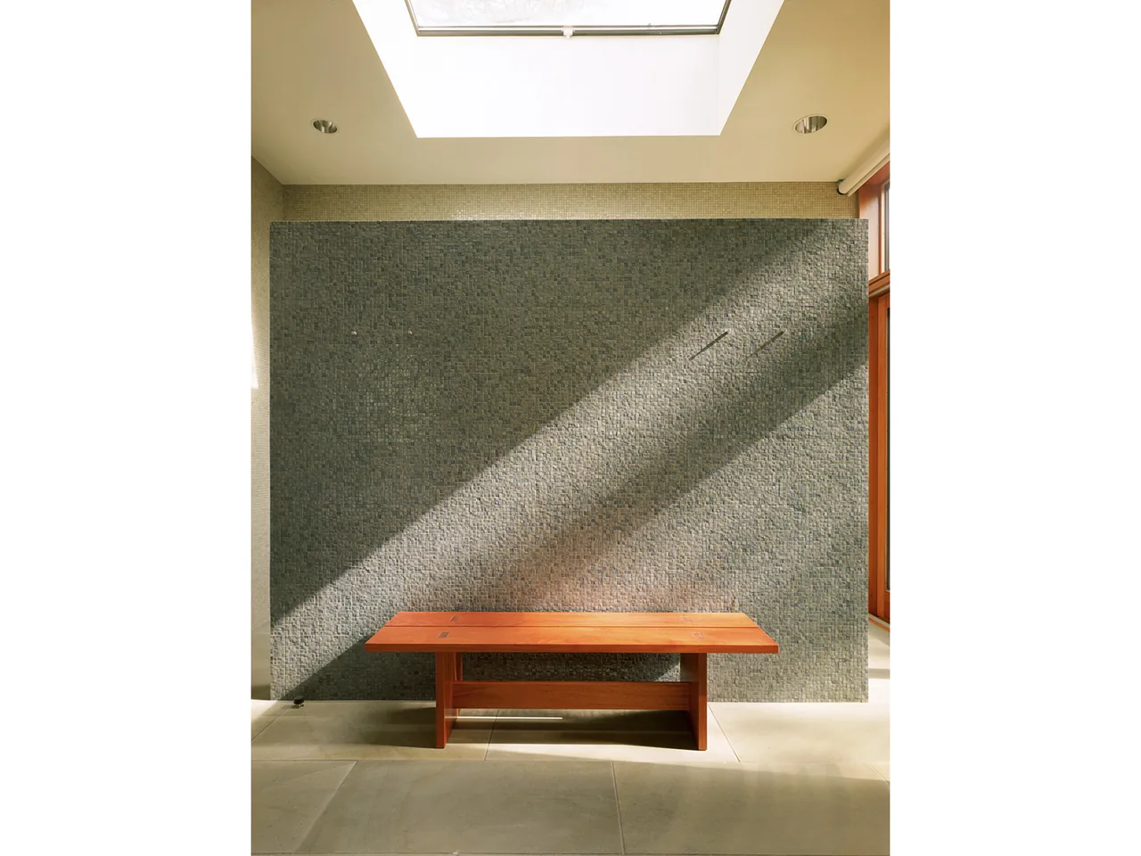 Wood bench in front of a gray stone tile wall illuminated by natural light from a ceiling skylight.