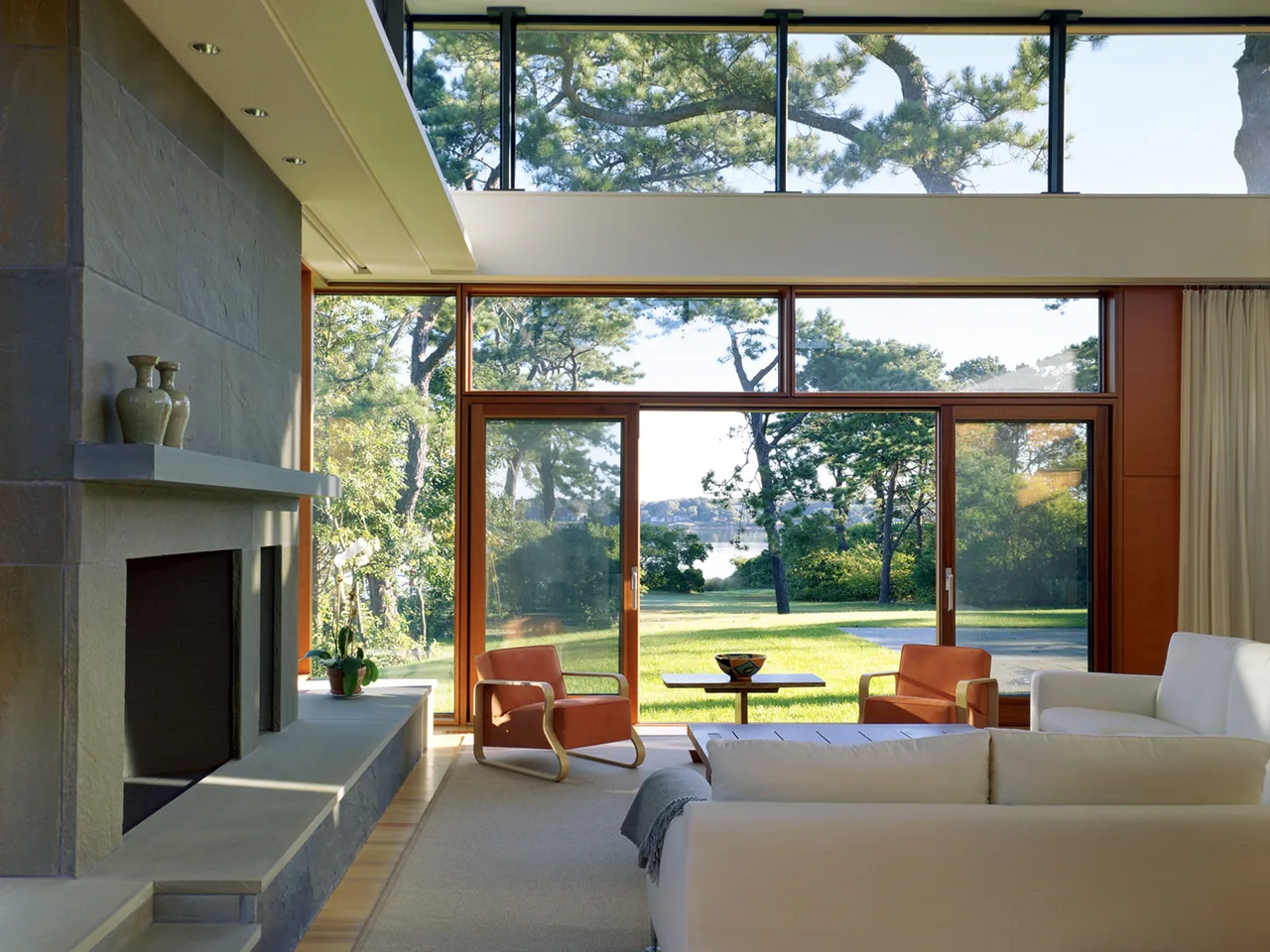 Living room in Long Island House features a large stone fireplace and expansive wood-framed glass doors and windows.