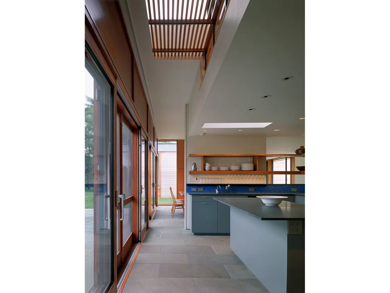 The kitchen in Long Island House features large sliding glass doors, open wood shelving, and a skylight above the island workspace.