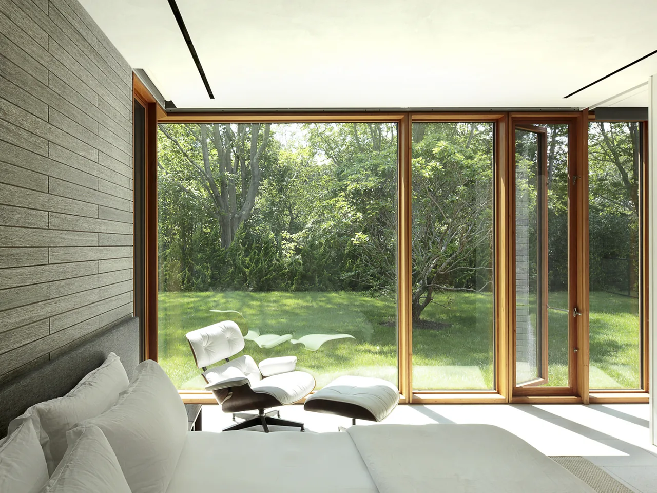 The Further Lane House bedroom features floor-to-ceiling wooden framed windows providing views of a green backyard.