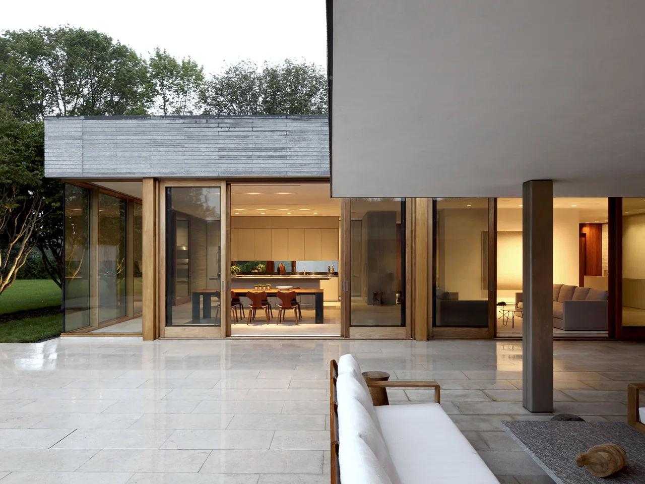 Modern house exterior with large wooden-framed glass doors opening to a kitchen and living room, set against a polished stone patio and greenery.