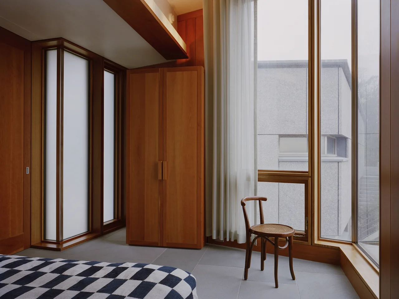 Bedroom corner with floor-to-ceiling glazing and wood cabinetry beside a woven seat chair.