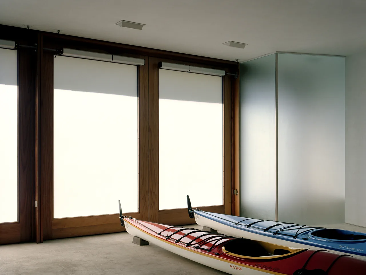 Two kayaks on a concrete floor in a room with wooden-framed windows covered by white roller shades.