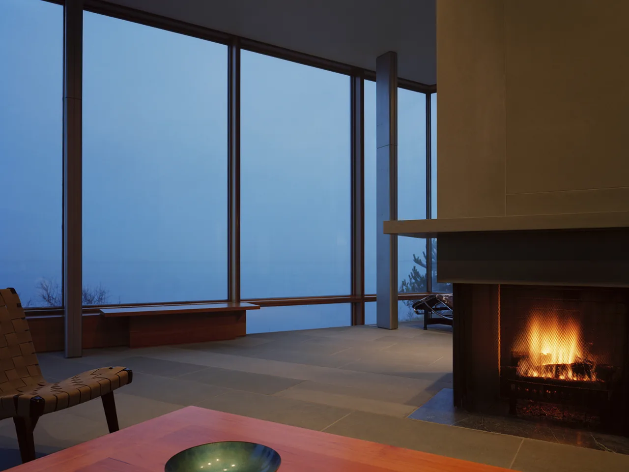 Shelter Island House living room features large floor-to-ceiling windows and a burning fireplace at dusk.