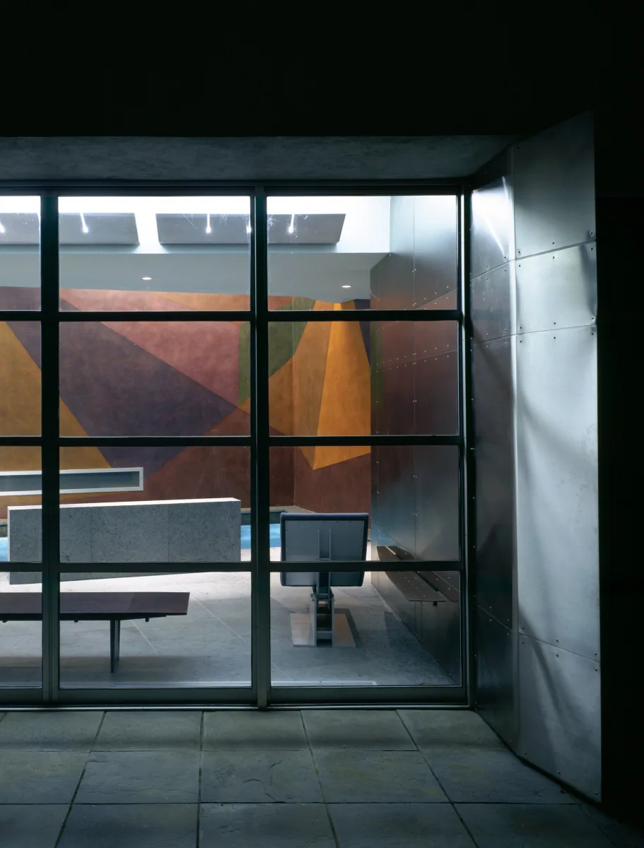 Indoor seating area viewed through a metal grid window highlighting an angular geometric mural inside the Pool House.