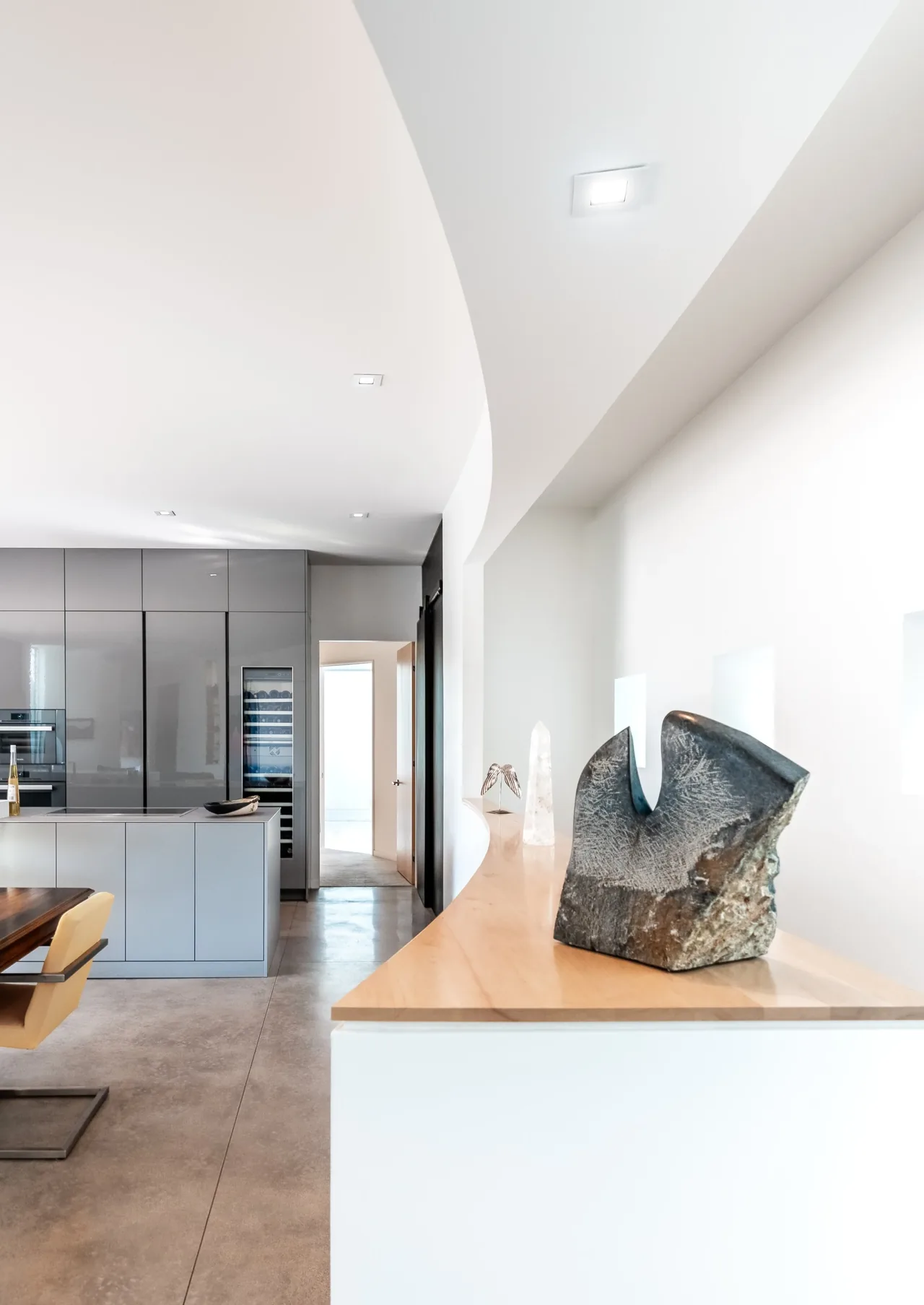 Modern kitchen with glossy grey cabinetry, a large island, and a curved wooden countertop displaying sculptures.