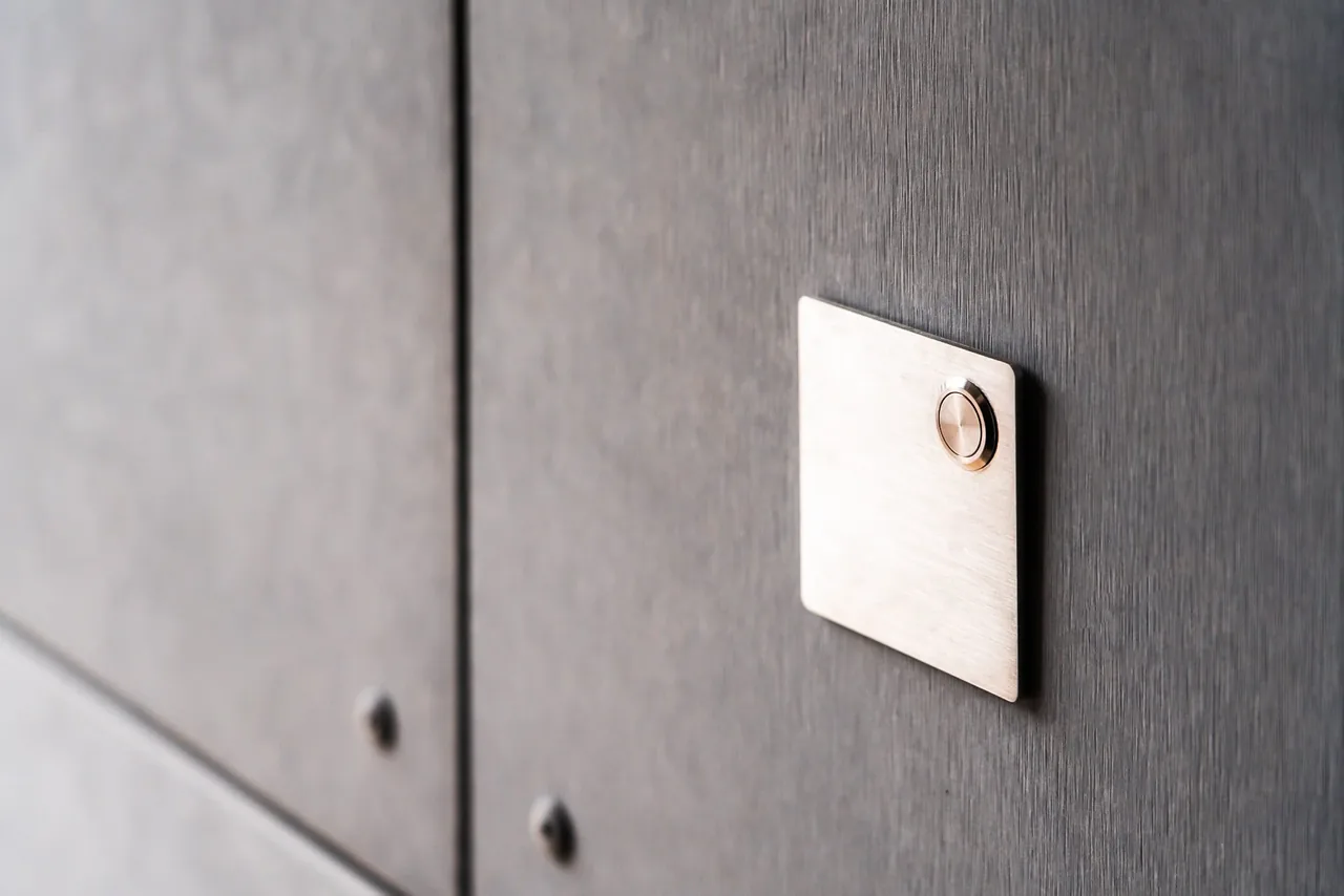 Minimalist metal doorbell button mounted on a dark textured wall at Jefferson St. Residence.