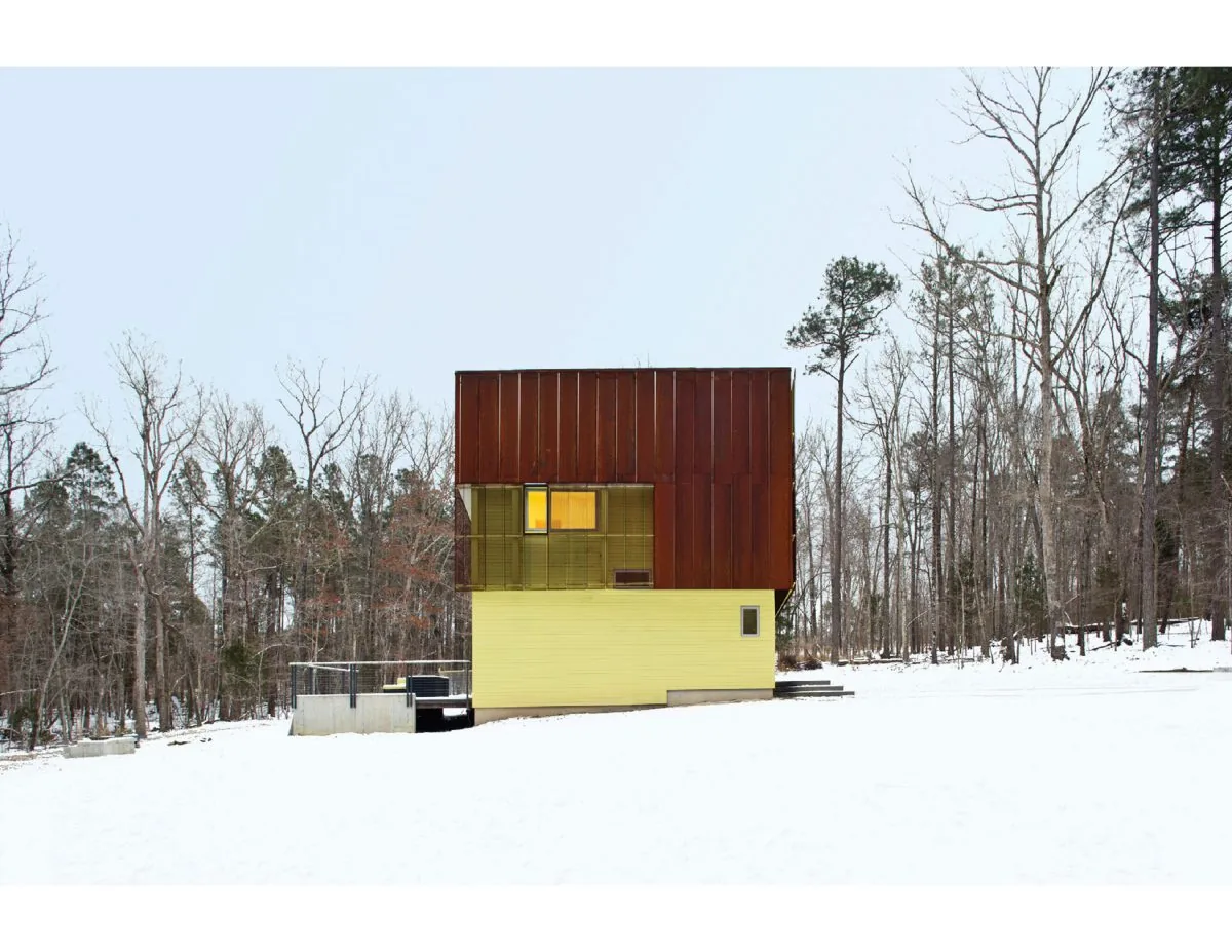 Weathering House showing layered yellow wood siding and perforated rusted metal panels in a snow-covered forest setting.