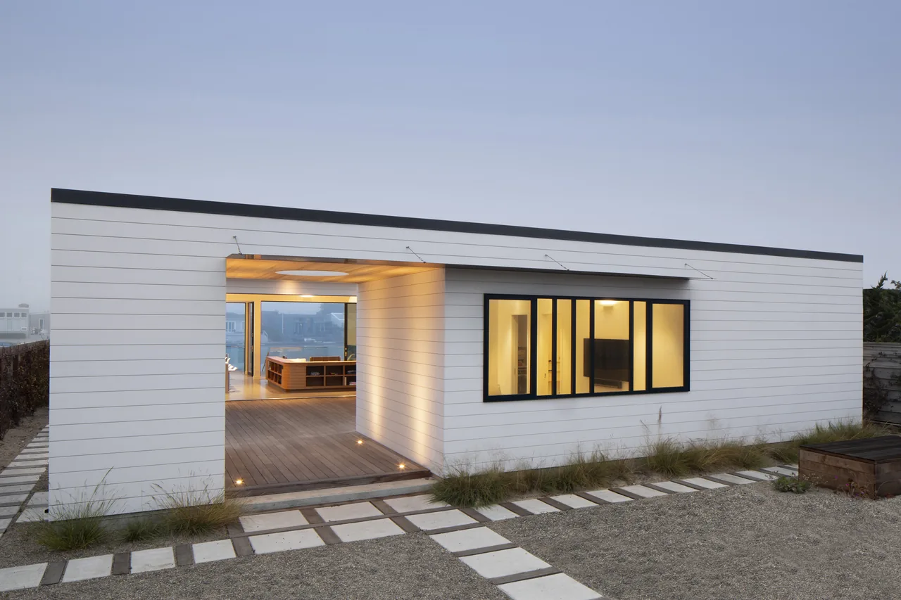 The Dune House by CCS Architecture features horizontal white siding, a flat roof, and a recessed outdoor living area with black window frames.