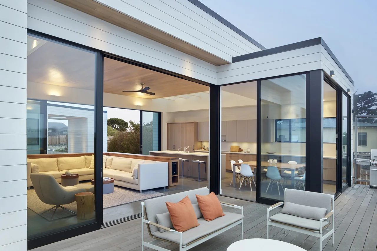 The Dune House by CCS Architecture features floor-to-ceiling black window frames with sliding glass doors connecting the deck to the living and kitchen spaces.