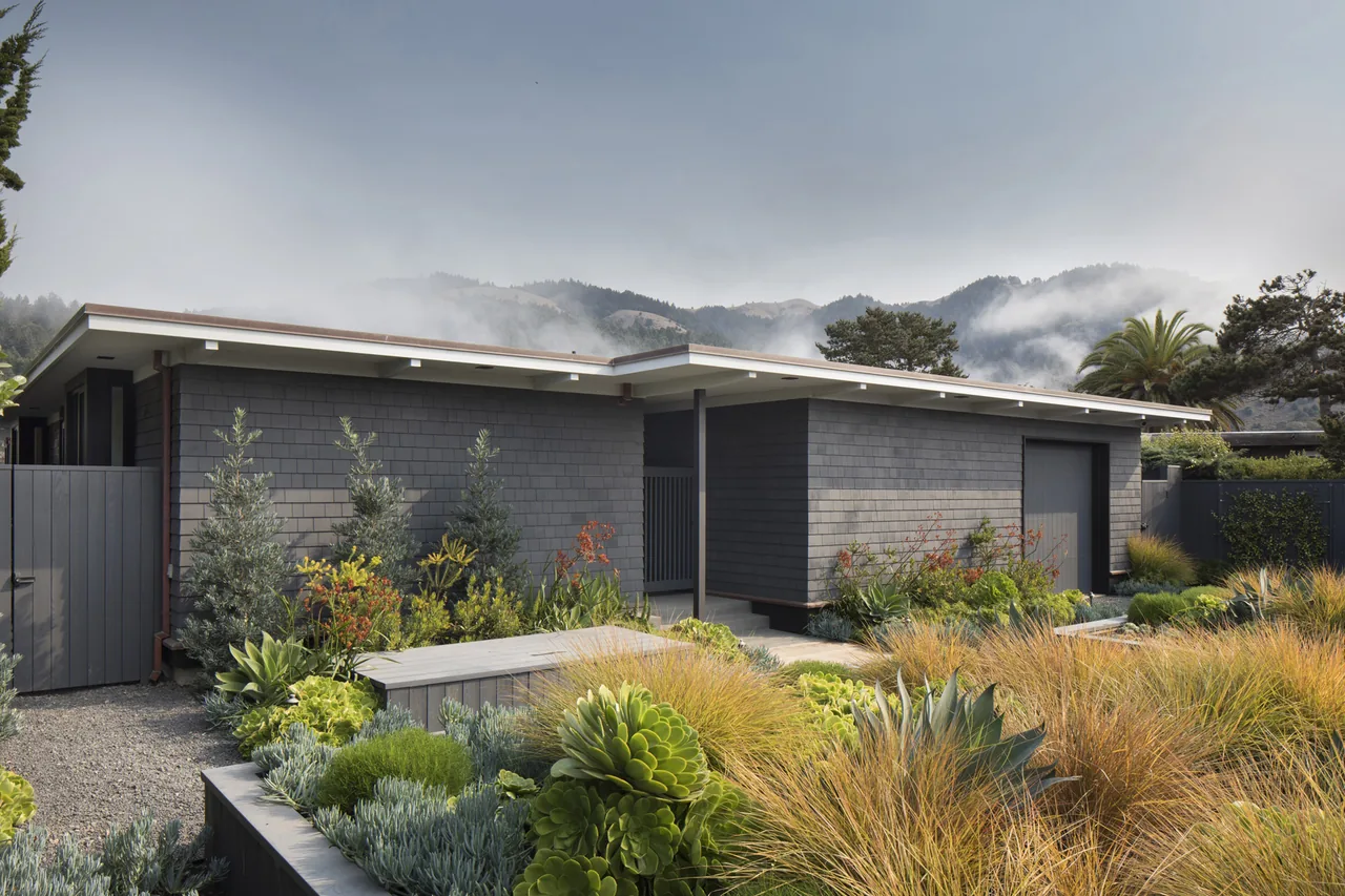 The Stinson Bungalow features dark shingle siding with a flat roof and a drought-tolerant garden in front of the house.