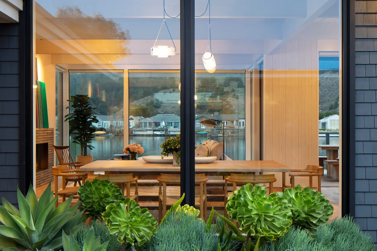 The Stinson Bungalow dining area features floor-to-ceiling windows overlooking a waterfront view with natural wood furnishings.