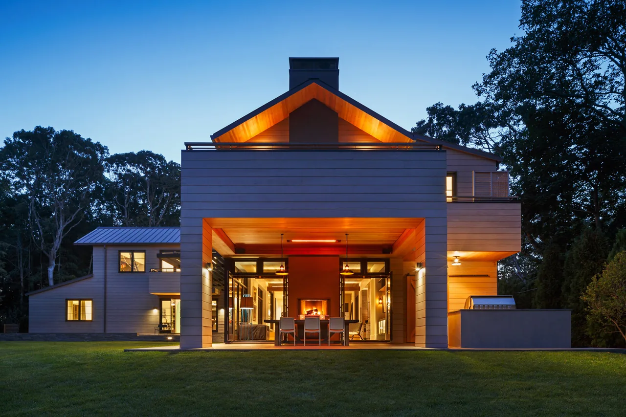 The rear facade of WATERMILL RESIDENCE features a covered patio with dining furniture and a fireplace, illuminated at dusk.