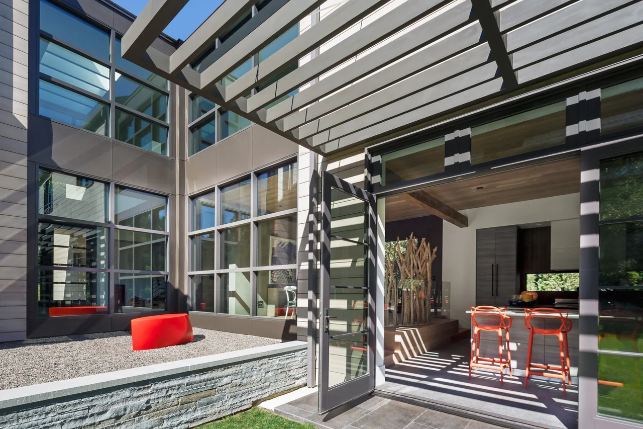 A modern outdoor living space with large glass windows and doors opening to a kitchen with vibrant orange bar stools, under a pergola.