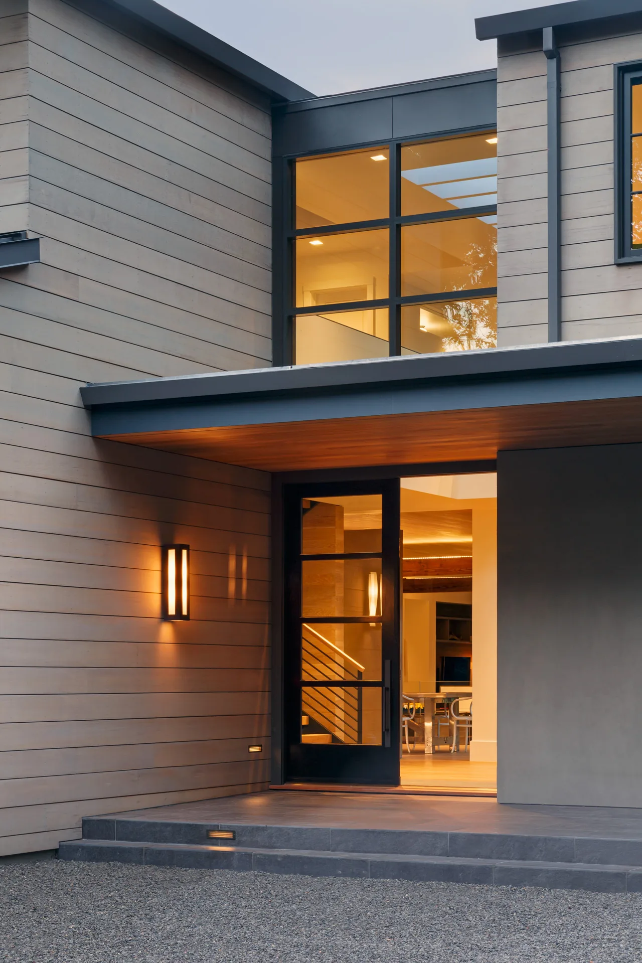 Modern home entrance featuring wood siding and black framed windows with warm exterior lighting.