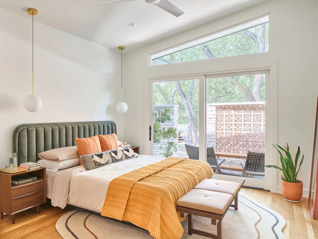 Bedroom with green velvet upholstered bed, orange accent pillows, and sliding glass doors leading to an outdoor patio with stone walls and trees.