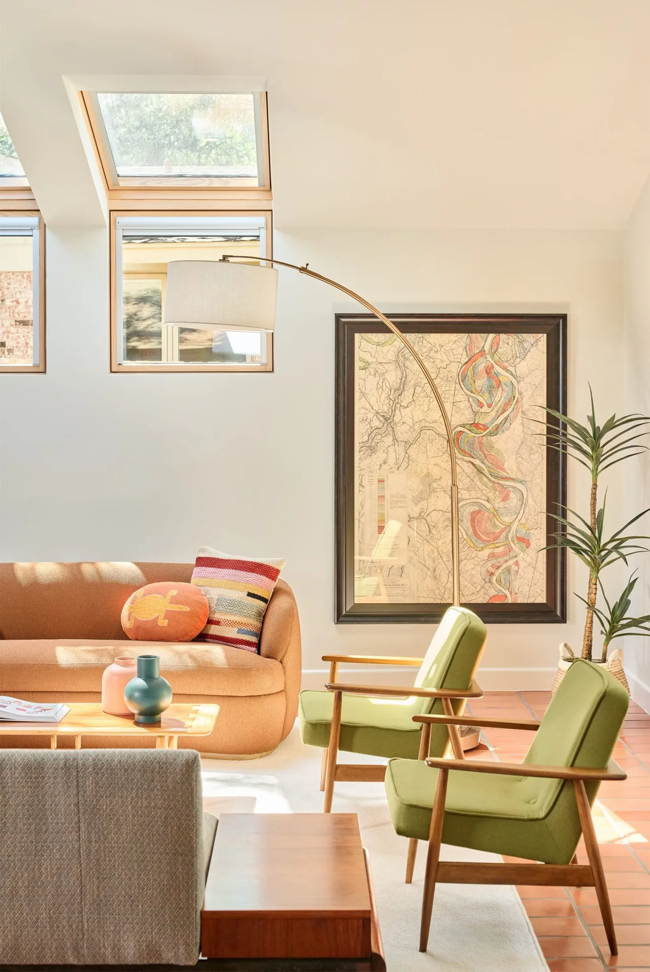 Daylit living room features midcentury modern green armchairs, an orange sofa, and a curved arc floor lamp beneath skylights.