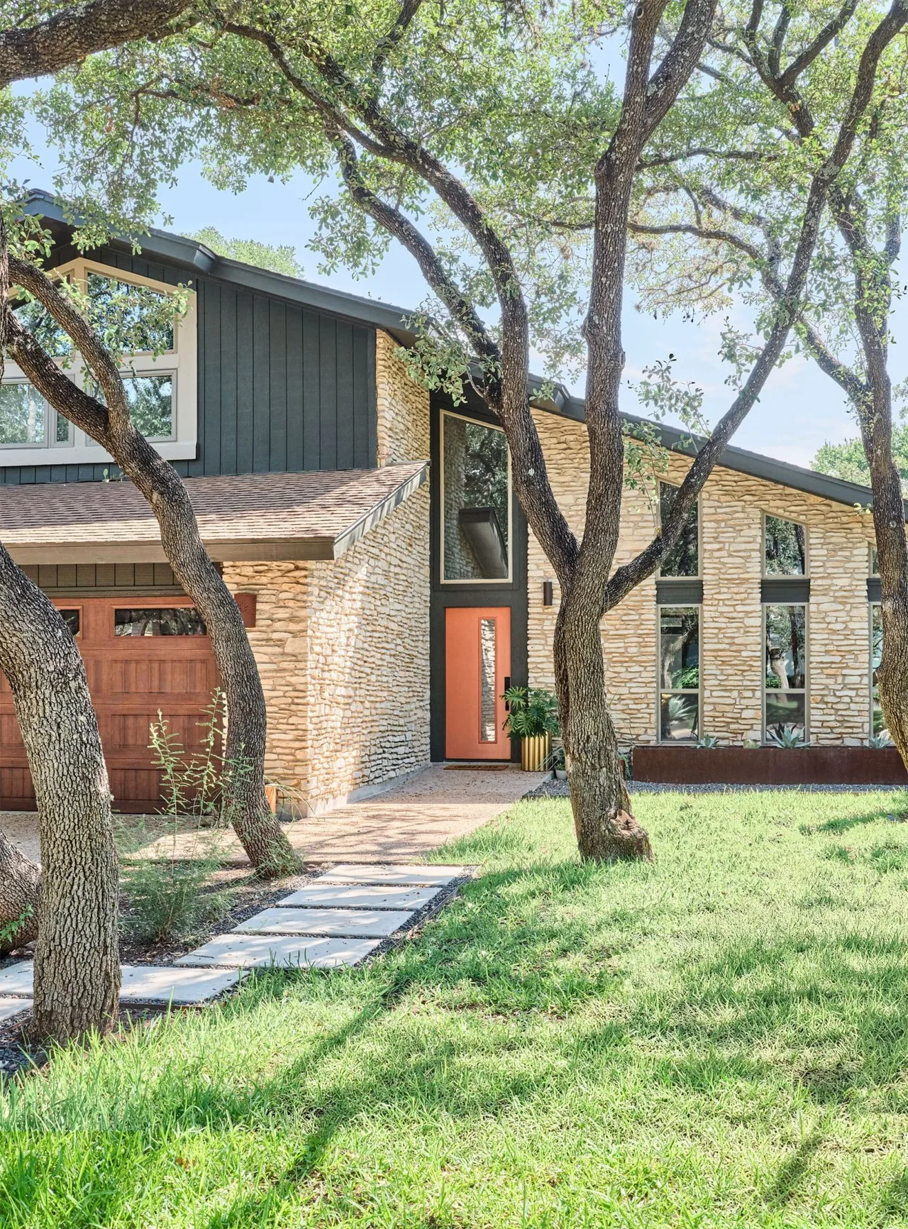 Modern residence named Settled In Light features stone siding, large windows, and a wood garage door surrounded by mature trees in Austin.