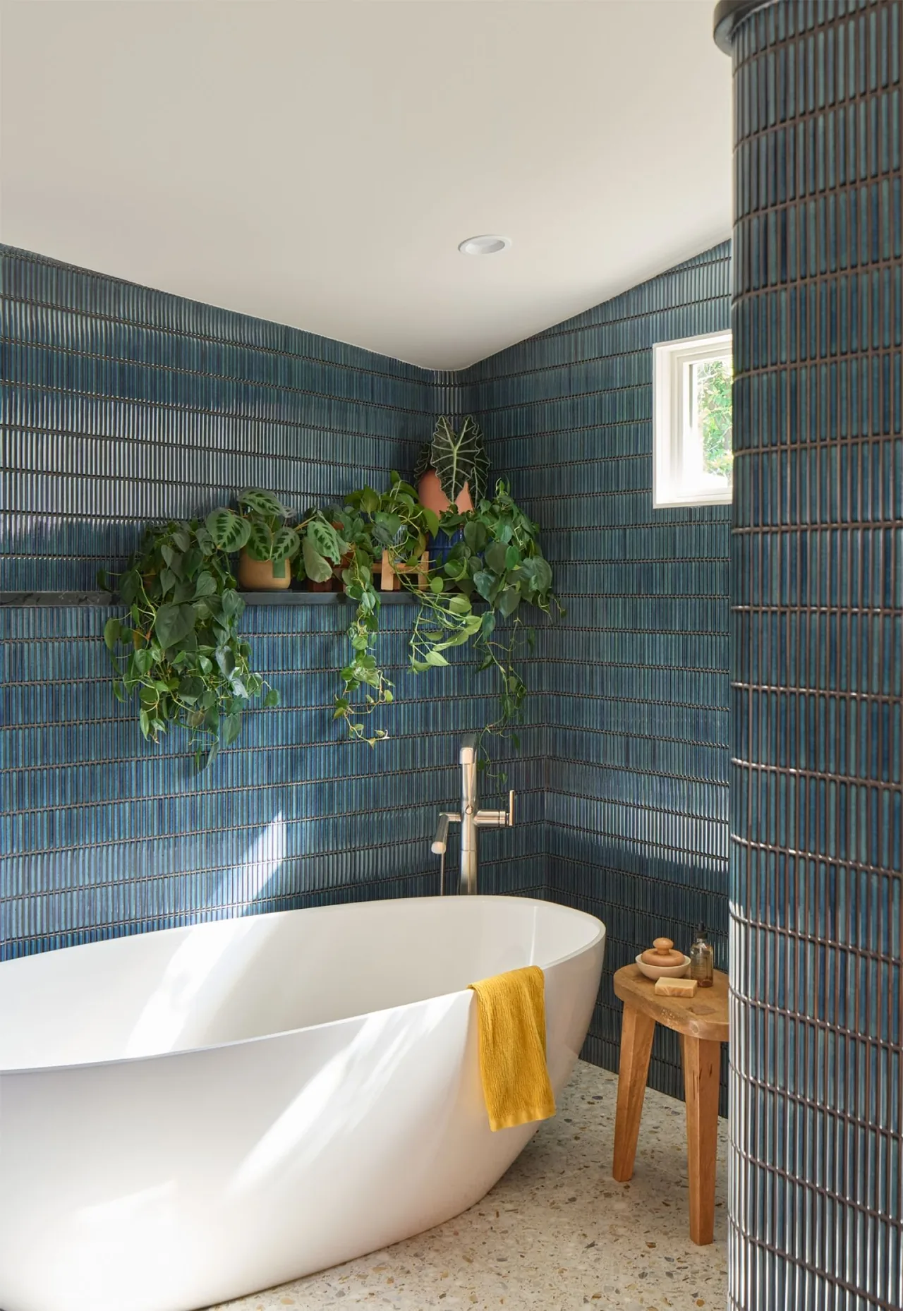 Bathroom with a white freestanding tub, blue vertical tile, and a wood stool with bath accessories.
