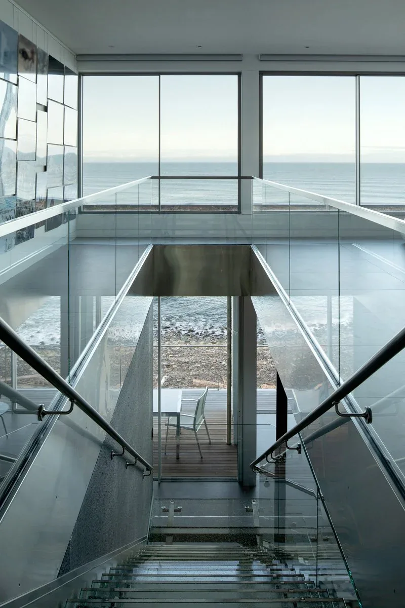 Glass-railed staircase descends to a deck with ocean views framed by floor-to-ceiling glazing in a modern house.