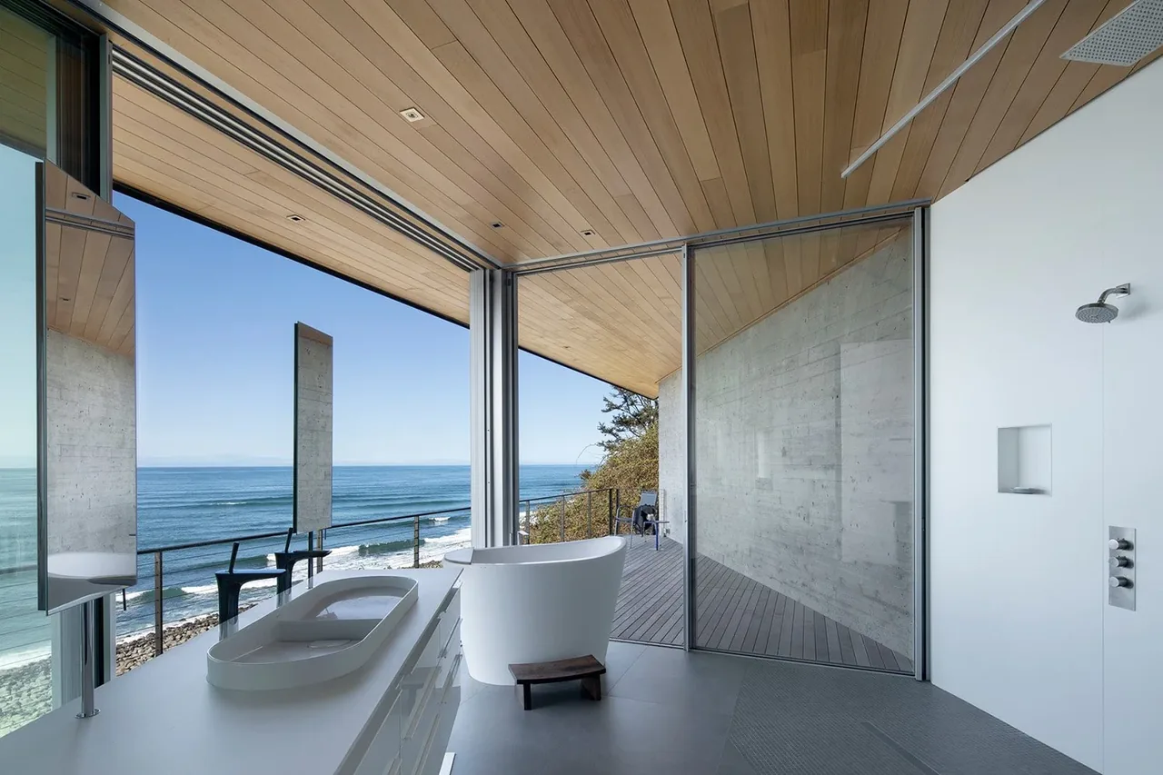 A bathroom features a freestanding tub with ocean views through large glass walls and a wood-paneled ceiling.