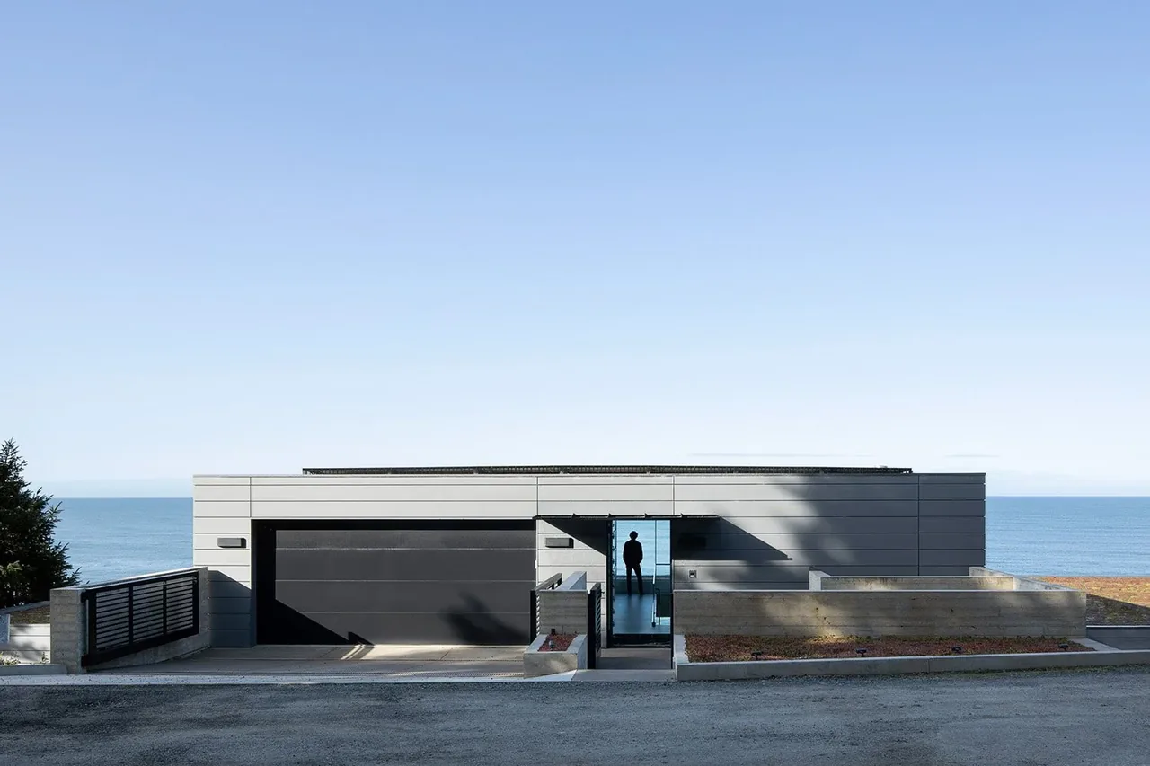 The modern prow house features a flat roof and reflective glass entry, positioned on a slope with ocean views.