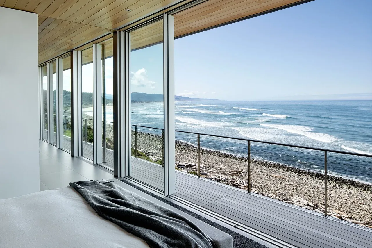 The prow house bedroom features expansive floor-to-ceiling glazing facing the ocean along a rocky shoreline.