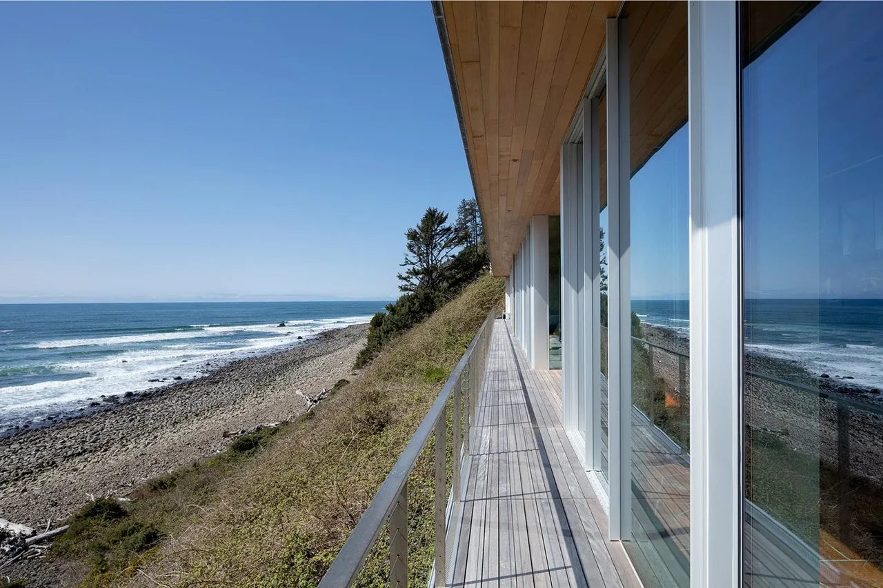 Exterior deck with metal railing provides ocean views along the side of a modern house near a rocky shoreline.
