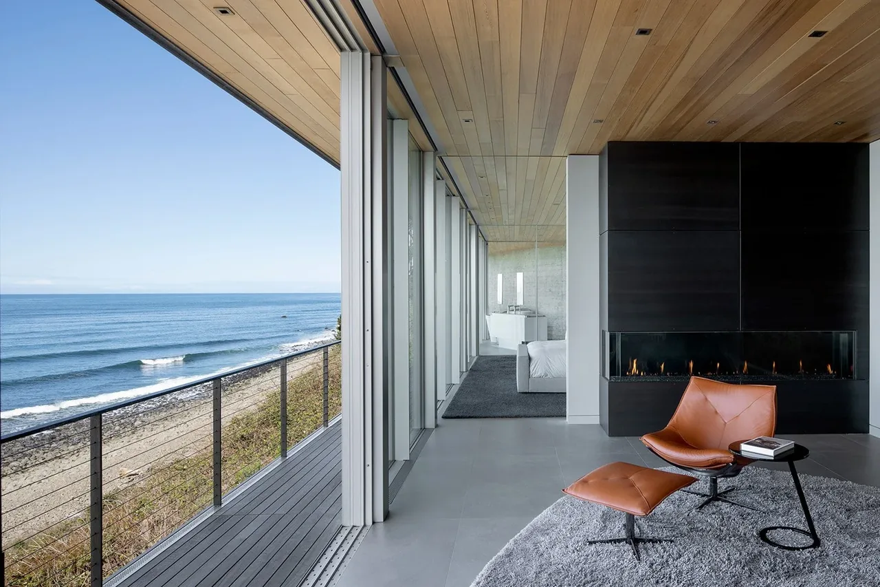 Living area of Prow House with floor-to-ceiling glass doors overlooking the ocean and a linear fireplace.