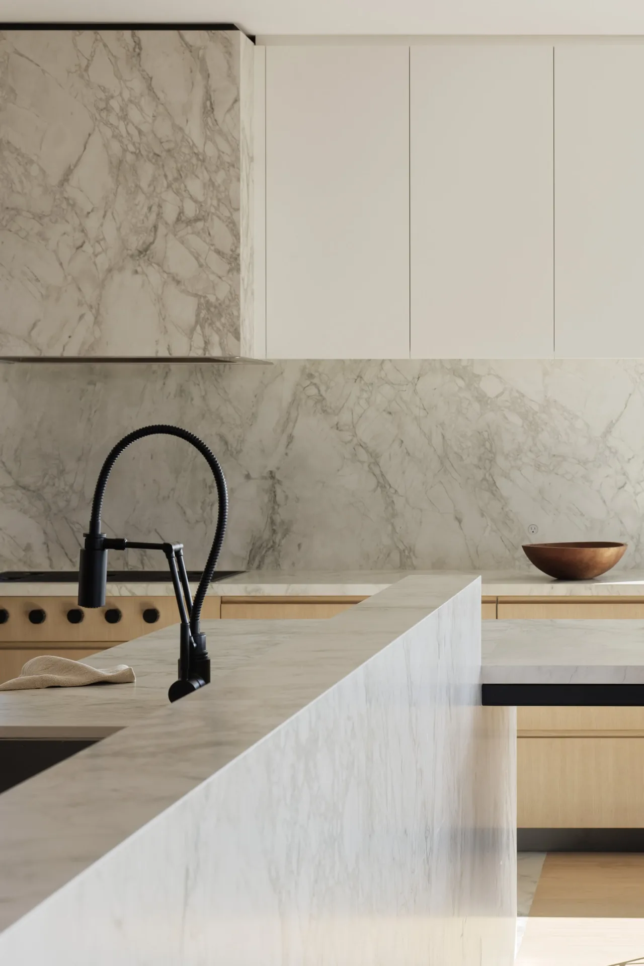 This kitchen features a marble peninsula and backsplash paired with minimalist white upper cabinets and light wood lower cabinets.