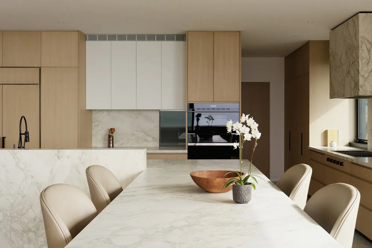 Modern kitchen featuring beige wood cabinets with marble countertops and backsplash, white upper cabinets, and built-in ovens.