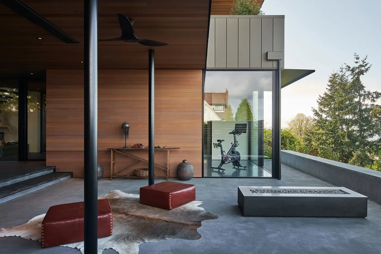 Concrete outdoor patio features leather poufs, a fire pit, and wood paneling, with a glass exercise room visible in the background.