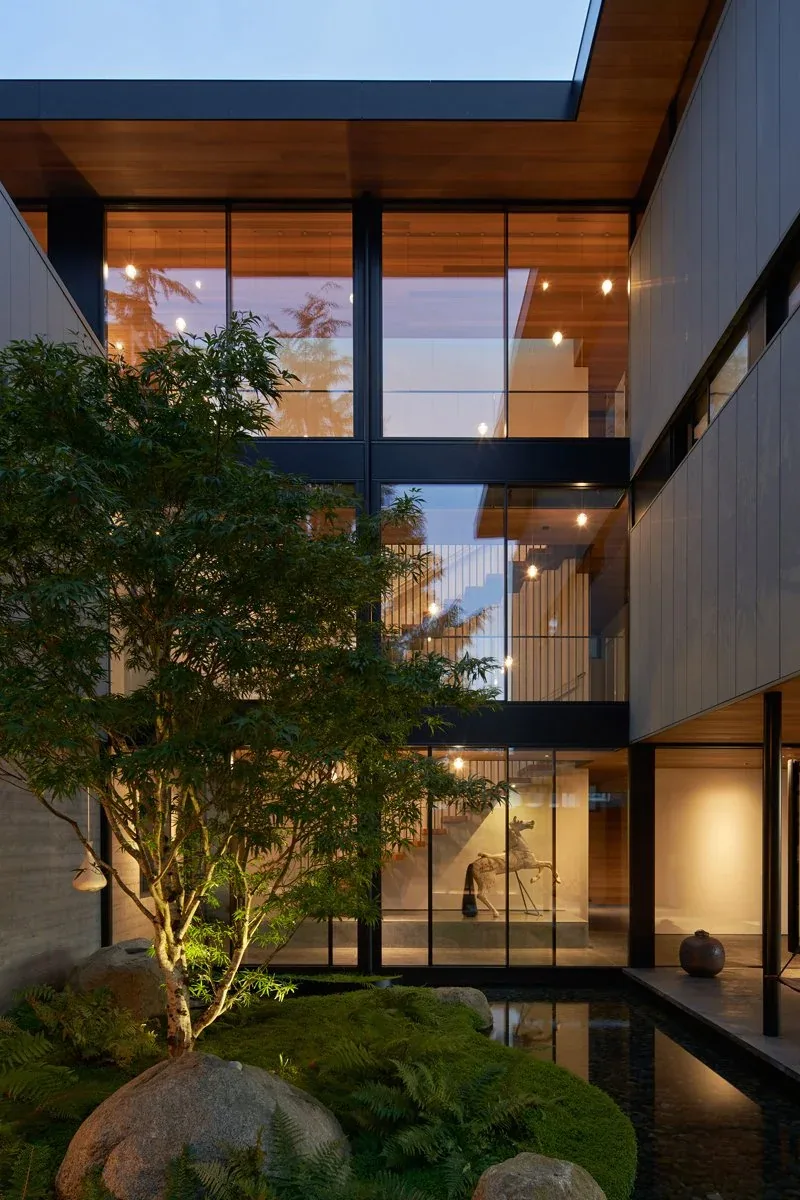 Multi-level modern courtyard features black window frames, wood soffits, and landscaped yard illuminated at evening.