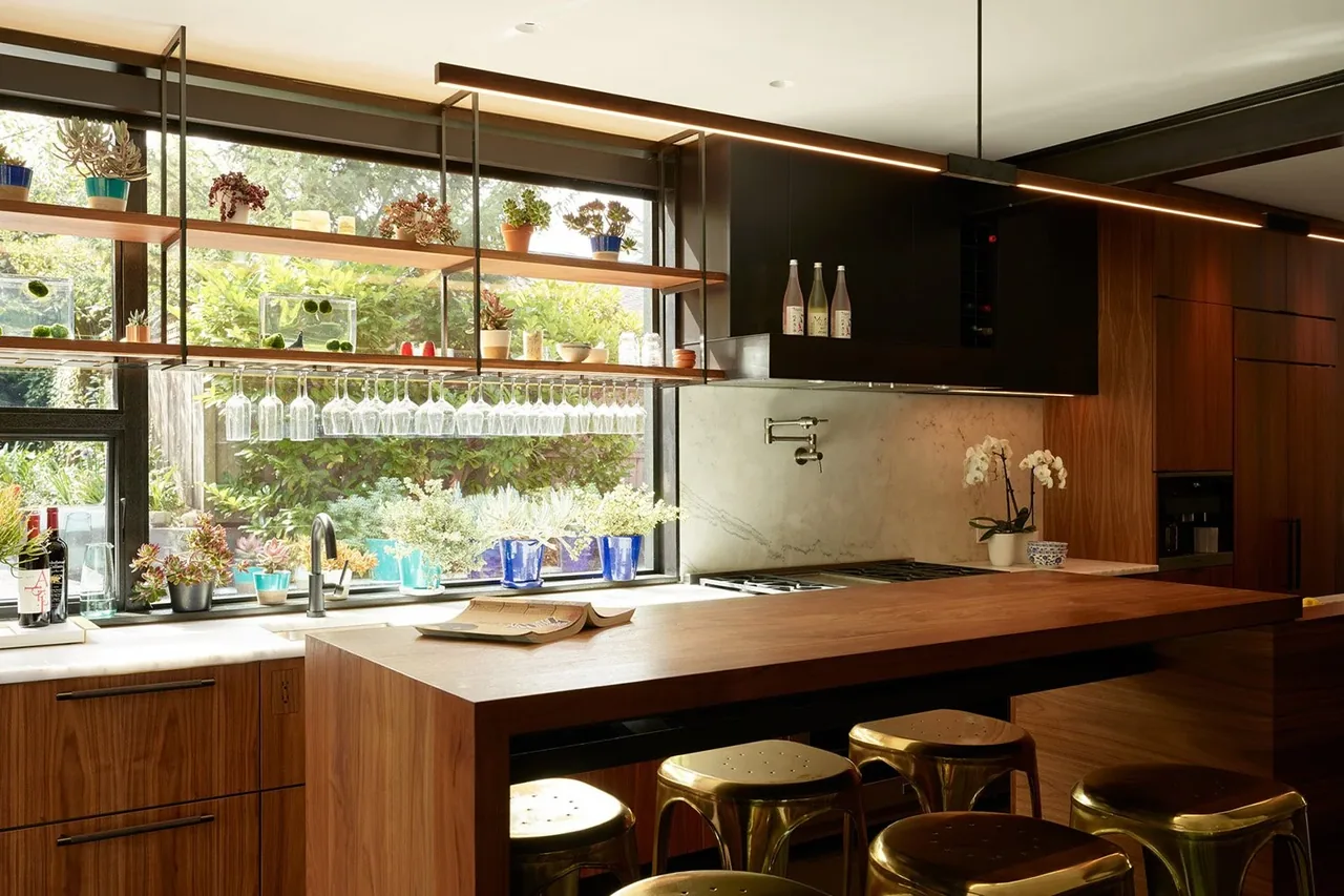 The Past Present House kitchen features wood cabinets, a marble backsplash, gold stools, and a large garden-facing window.