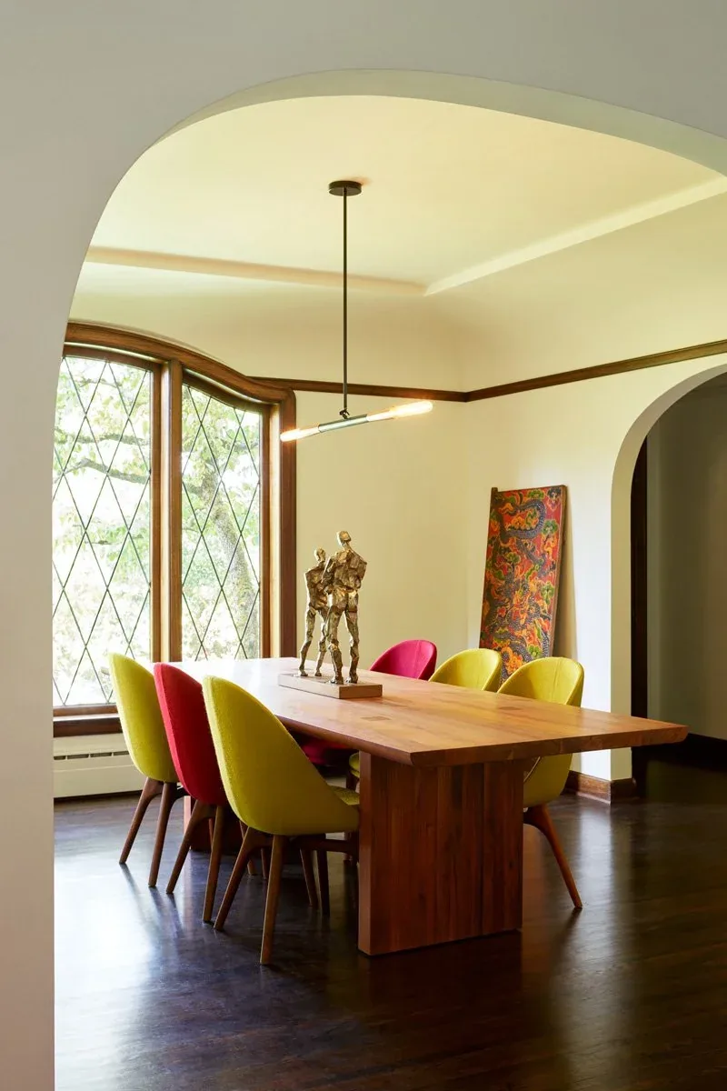 A wood dining table surrounded by yellow and red chairs sits beneath a modern pendant light by leaded glass windows.
