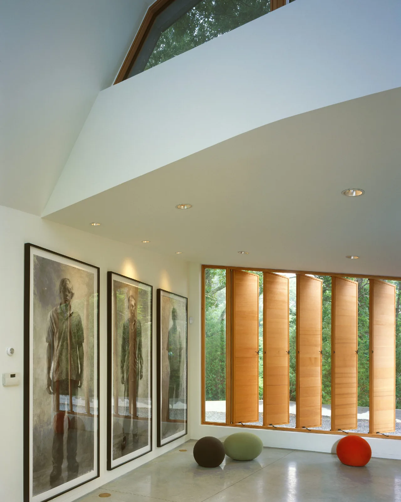 Room corner with vertical wooden window panels, three framed portraits, and spherical cushions on a polished concrete floor in Orleans House.