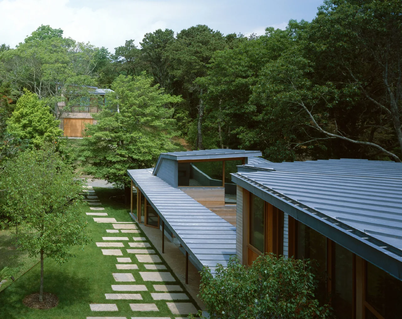 Orleans House in Cape Cod features a standing seam metal roof and a stone path through a green lawn, blending with wooded surroundings.